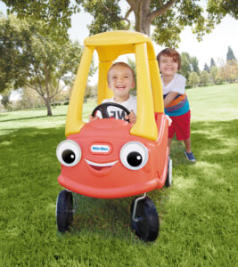 A photo of two young boys smiling and laughing as they push and drive around the little tikes cozy coupe in the park